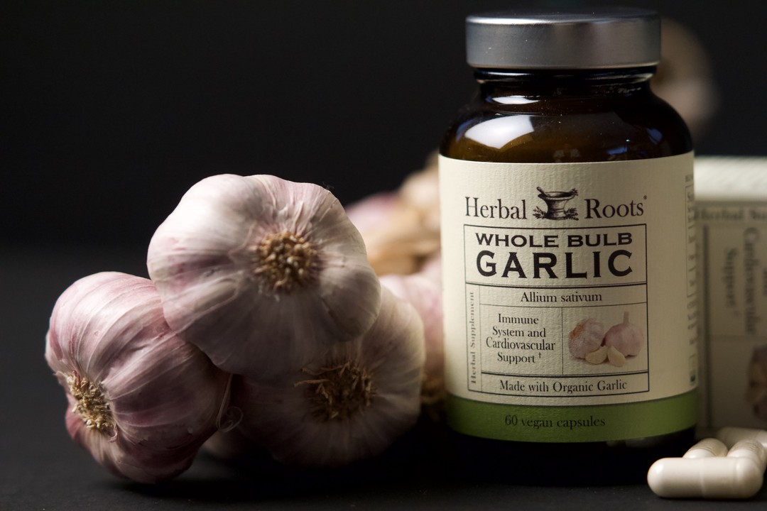 Bottle of Herbal Roots Organic Garlic with capsules on the right of the bottle and three garlic cloves on the left.