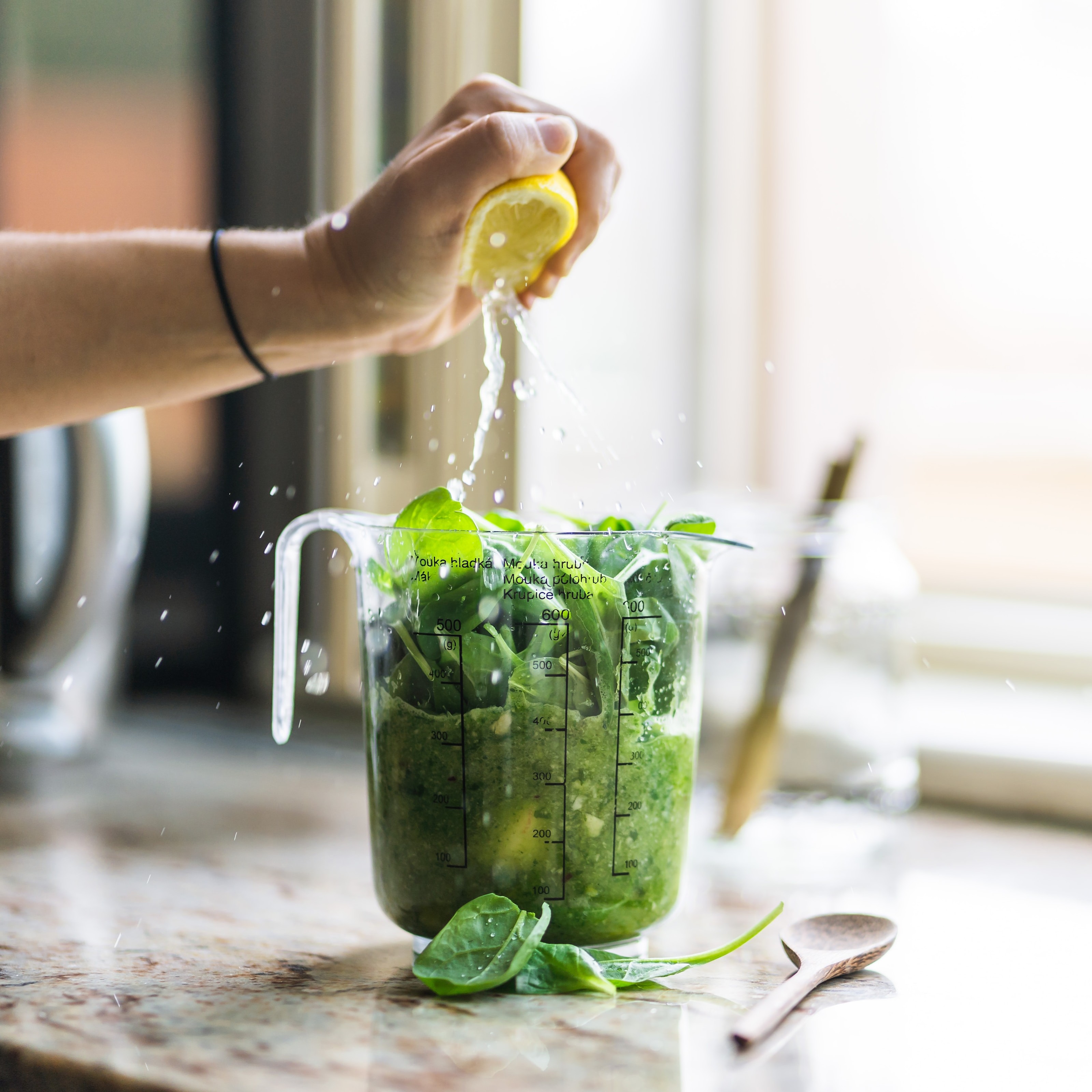 A hand squeezing half a lemon over a measuring cup full of leafy greens set on a counter with a wooden spoon beside it.