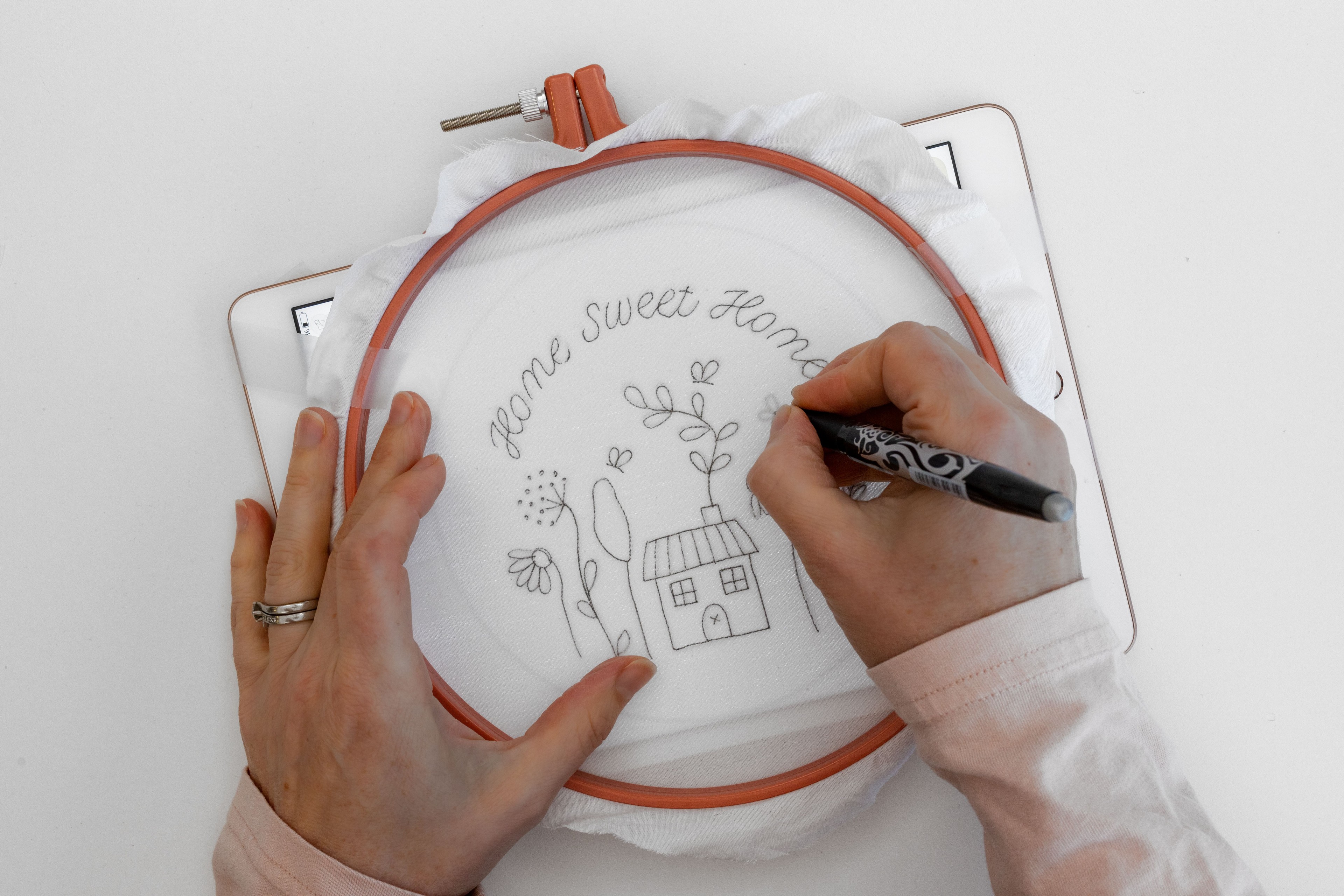 A hand draws a floral Home Sweet Home on the fabric with the Ipad behind it.