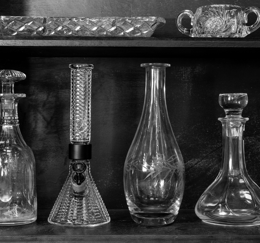 Black and white lifestyle photo of the clear Prism x Jane West Twenties MINI Beaker Bong, placed among vintage whiskey decanters and etched vases. The image evokes the 1920s Prohibition era, emphasizing the Art Deco diamond texture as a symbol of rebellion and refined taste during the 2020s cannabis prohibition era.