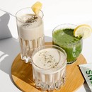 Three glasses of smoothies - one smoothie in a tall glass containing a beige colored smoothie with chia seeds on top and a pineapple wedge. The smaller glass has a greens smoothie and a lemon wedge. The smaller glass that is in front has a beige colored smoothie with seeds on top. Image set against white background. glasses are on a small wooden lazy susan.