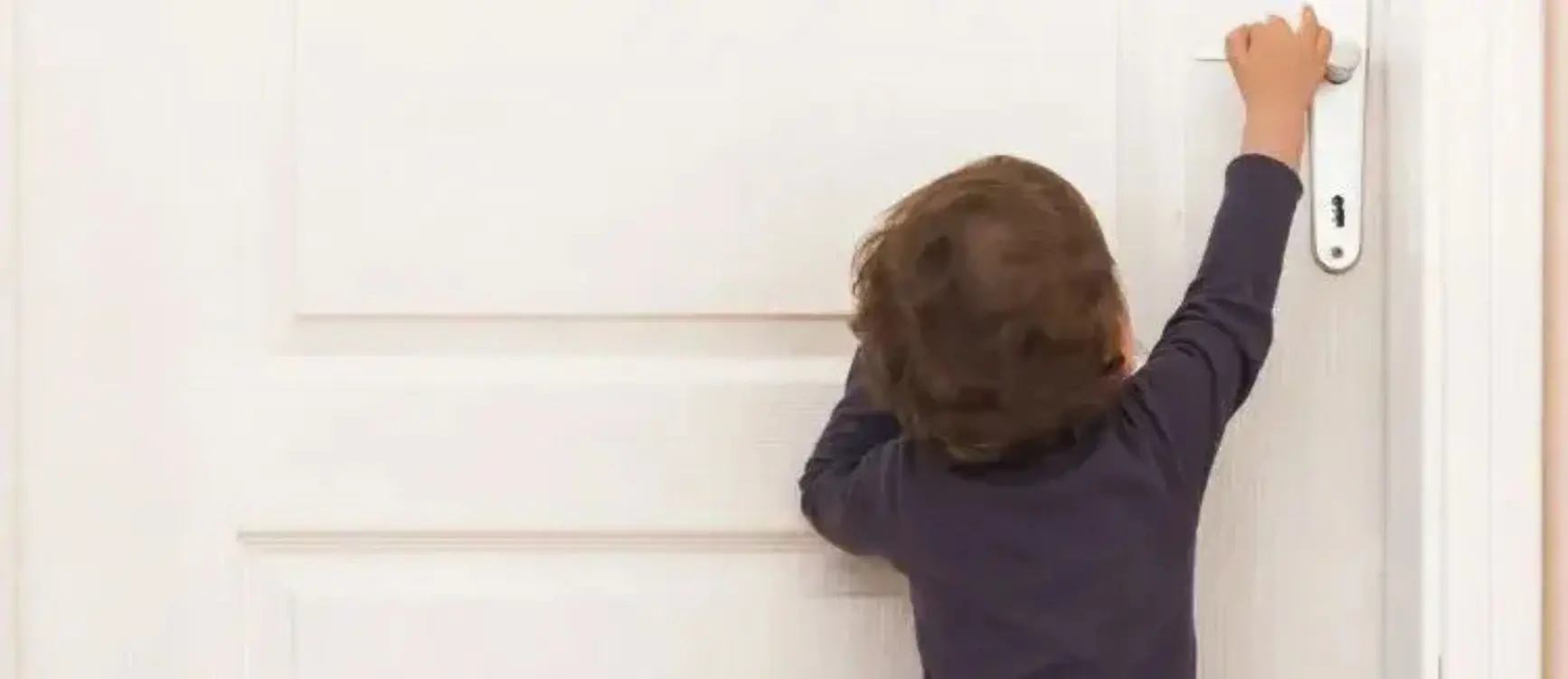 A young boy stretches his arm to grasp the handle of a door, showing curiosity and eagerness to enter.