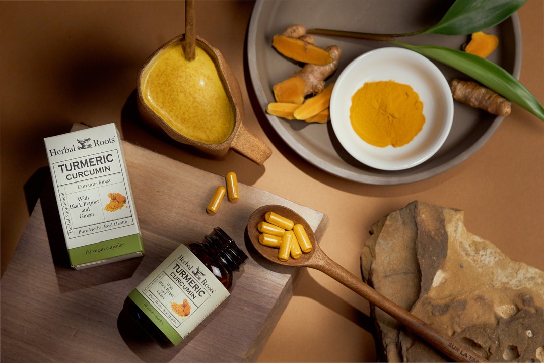 aerial view of a bottle of herbal roots turmeric supplement on its side with a cutting board next to it that has a wooden spoon with turmeric capsules in it and a mug of orange drink. To the left of the bottle is a ceramic plate with a small bowl of turmeric powder in it and a small pile of chopped turmeric root in front of it.