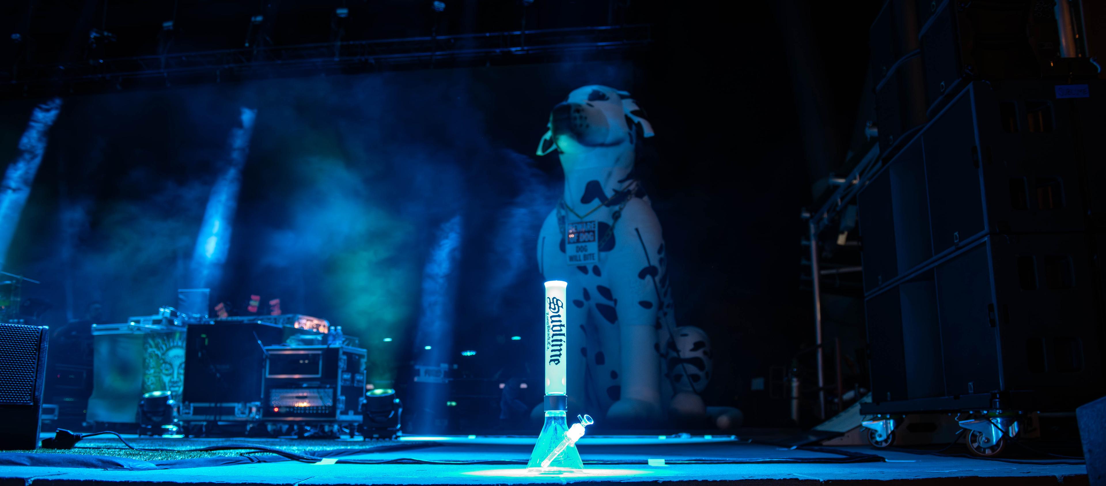 A tall white Sublime x Prism official modular glass bong illuminated by stage lights at Red Rocks Amphitheatre. The piece is positioned near a large inflatable Lou Dog dalmatian during the historic 2026 show where Jakob Nowell was officially inducted as the new frontman of Sublime.