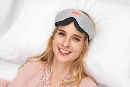 A smiling blonde girl wearing a contoured sleep mask on her head while lying down on a white pillow.