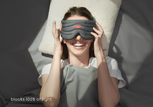 A smiling girl lying down on a pillow while holding the edges of a weighted sleep mask.