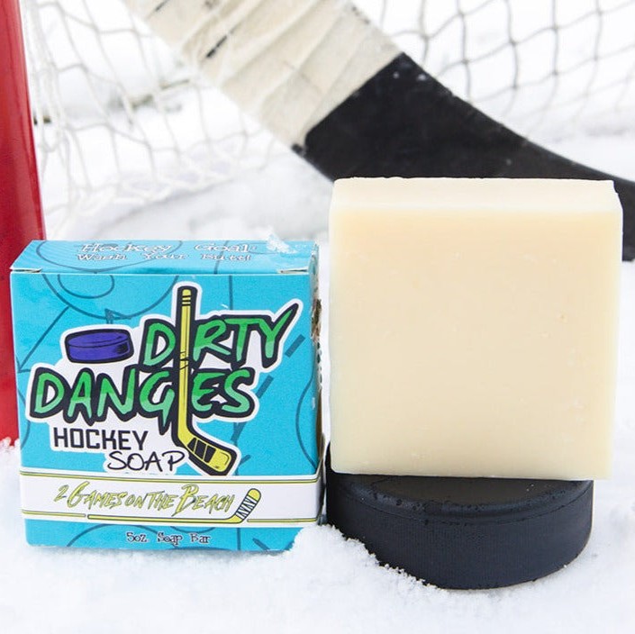 A yellow bar of dirty dangles hockey soap sits on a hockey puck in the snow with a hockey stick and a hockey goal. 2 Games on the Beach scent.