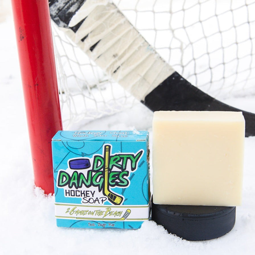 a yellow hockey soap bar sits on a hockey puck in the snow with a hockey stick and a blue soap box. Dirty dangles Hockey Soap 2 games on the beach scent.