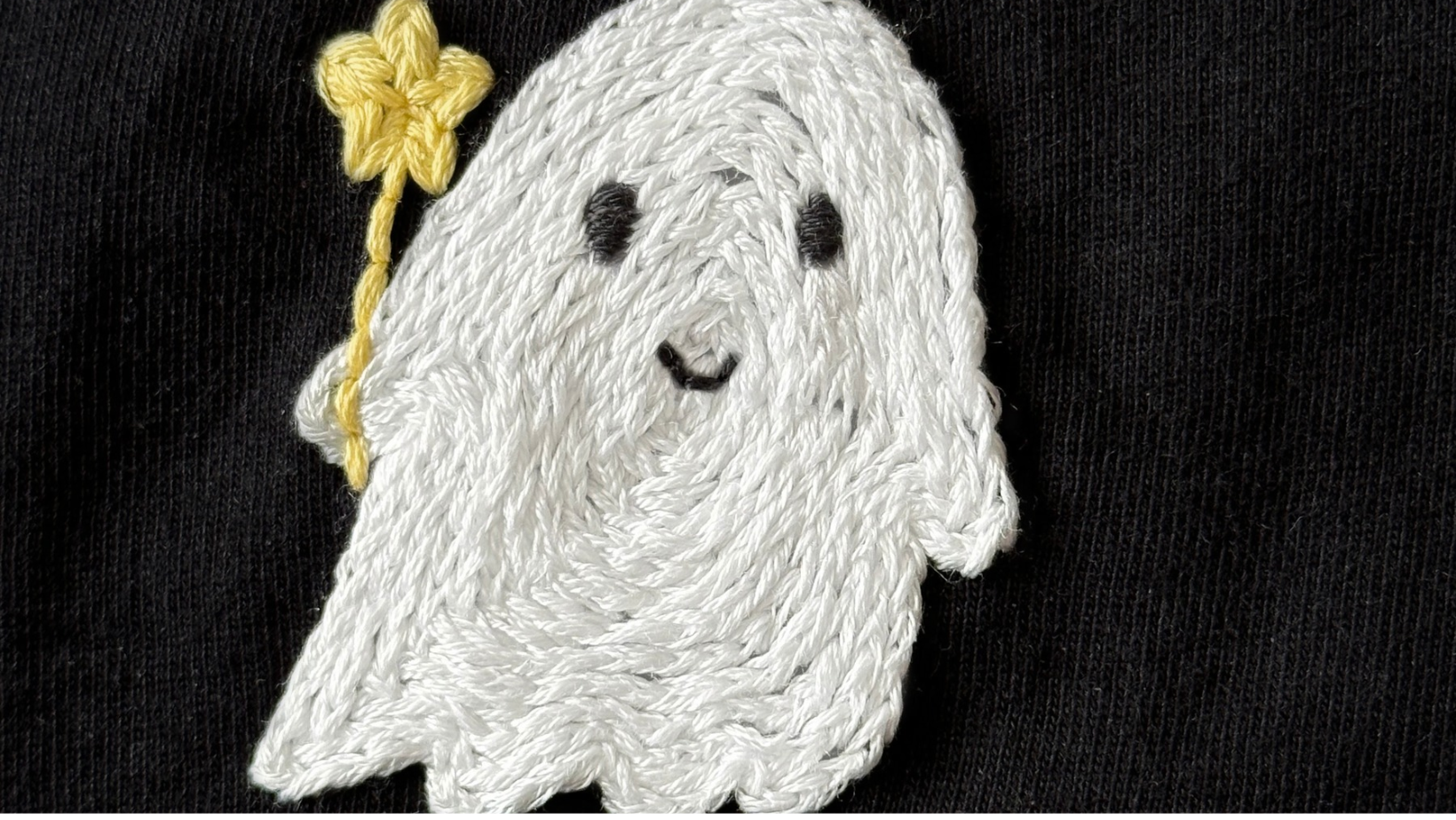 A close-up of a white ghost embroidered on black fabric, showing the rows of stitching.
