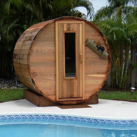 Image of an outdoor home sauna next to a pool. It is a classic barrel shape with a window door.