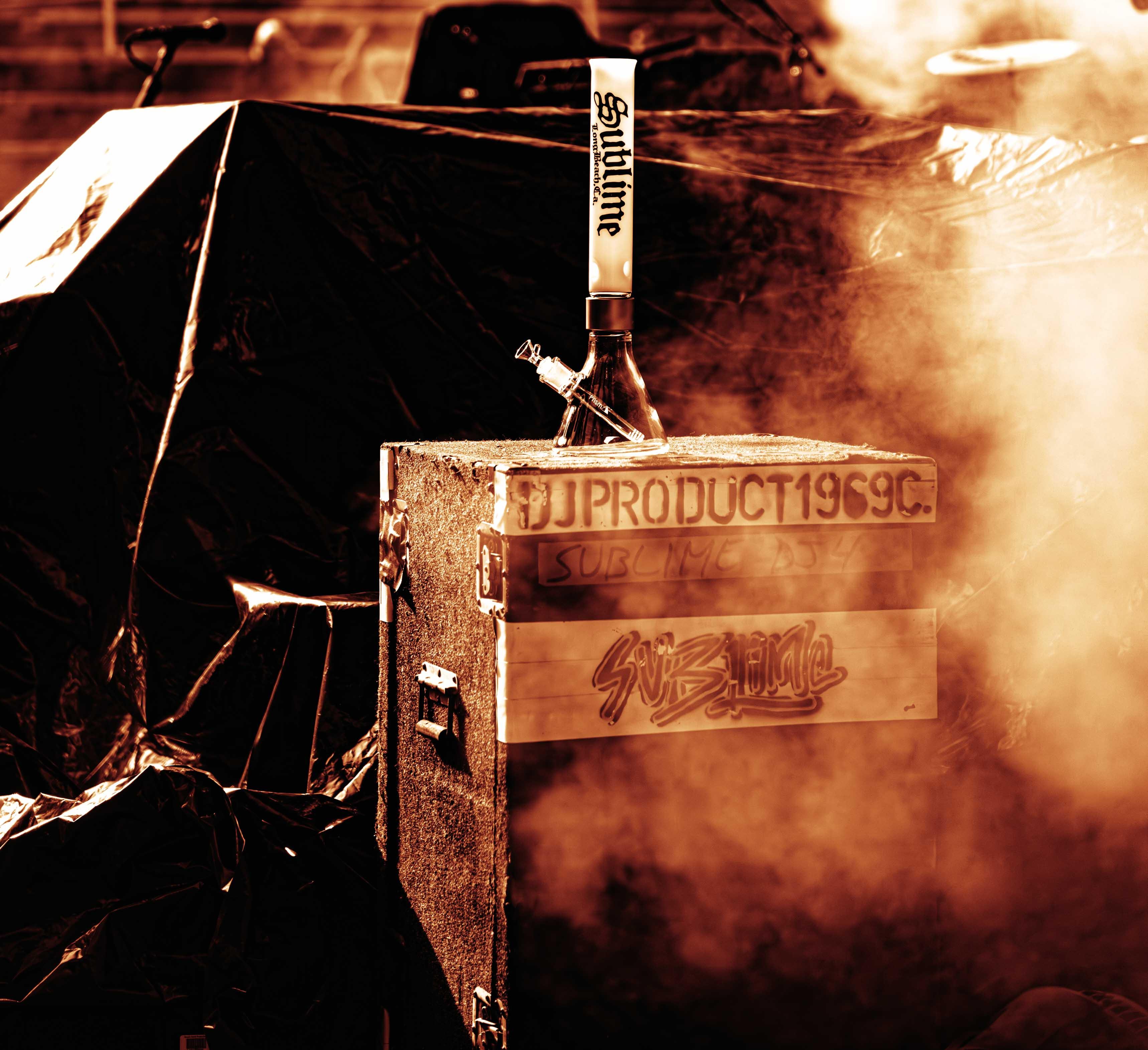 A tall white Sublime x Prism official modular glass bong sits on the Sublime crew’s equipment cases backstage at Red Rocks Amphitheatre. Surrounded by a cloud of smoke, the piece is shown with the band’s tour gear ahead of their 2026 performance.