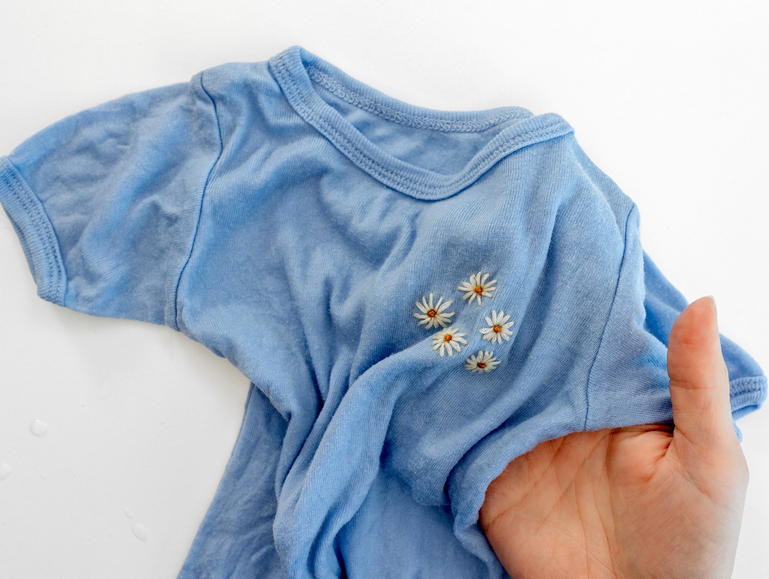 A hand holds a shirt up showing the embroidered daisy design.