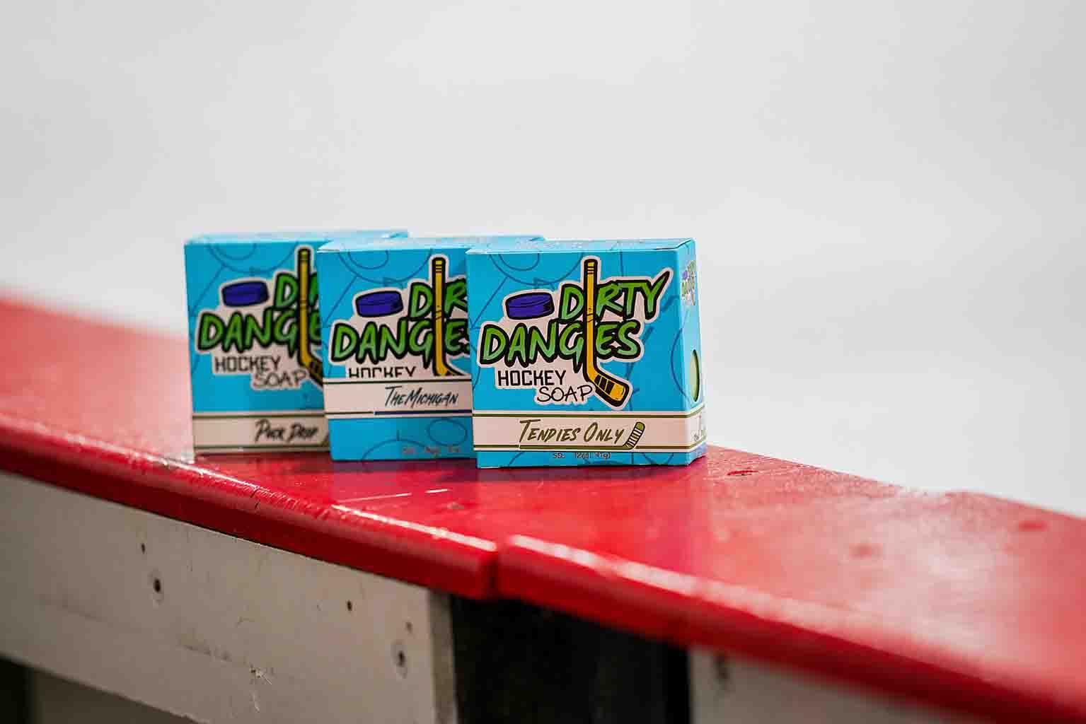 Three Dirty Dangles hockey soap boxes—Puck Drop, The Michigan, and Tendies Only—lined up on a red rink bench.
