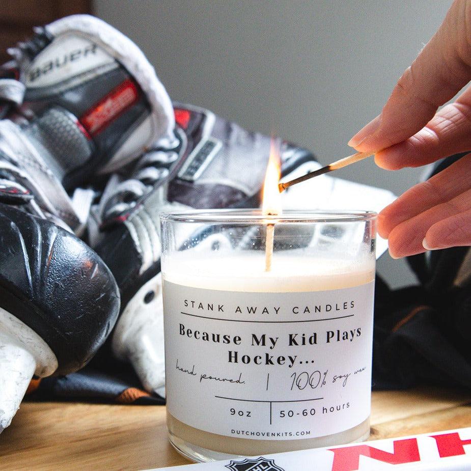 A white hockey candle sits on a table with a hockey stick and a hockey helmet. A hand lights the candle with a match. Because my kid plays hockey