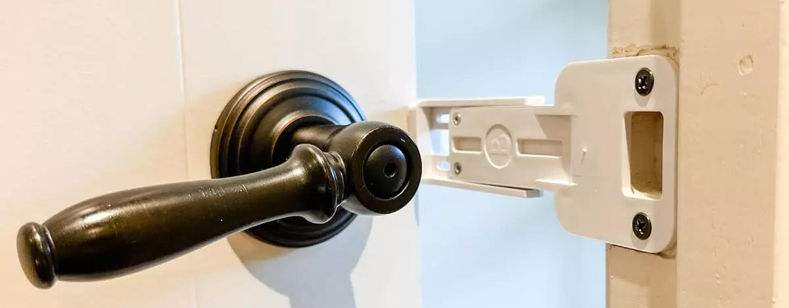 Close up of The Door Buddy door prop for cats installed between a door handle and the strike plate on the door frame. The prop replaces the strike plate and holds the door slightly open to create a cat sized opening while helping keep dogs out.