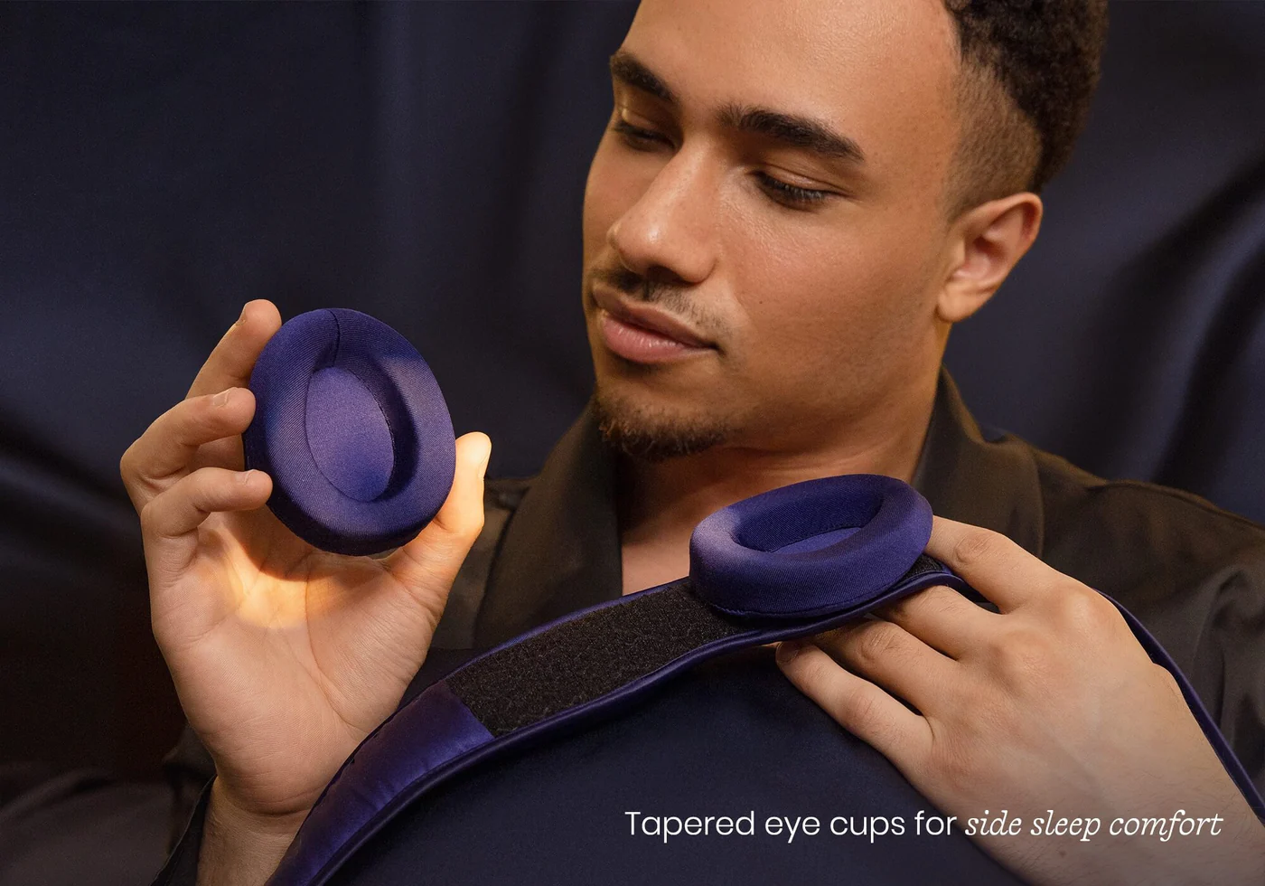 A man holding a convex eye cup in one hand. And with the other hand, a head strap of an adjustable sleep mask with another eye cup attached.