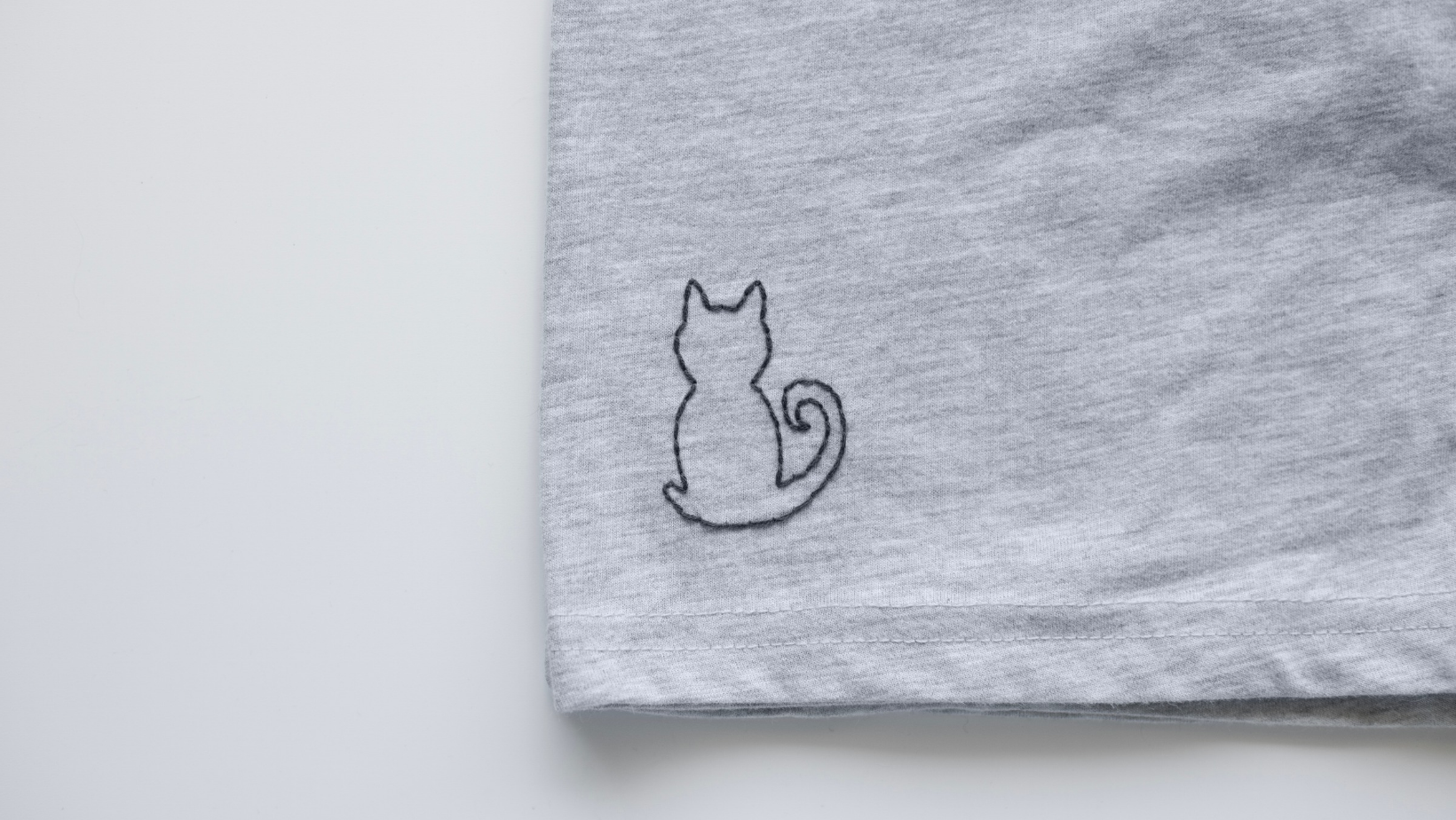 cat outline embroidered in black thread on the hem of a grey t-shirt.