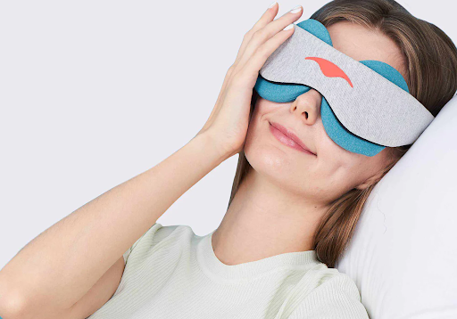 A girl lying down on a pillow touching the side of the gray cooling therapeutic eye mask with blue eye cups that she’s wearing.