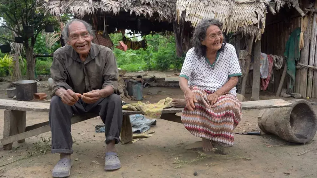 Elderly Indigenous man and woman from Tsimane tribe sitting on wooden benches outside a thatched-roof hut, smiling and appearing relaxed in a rural setting.