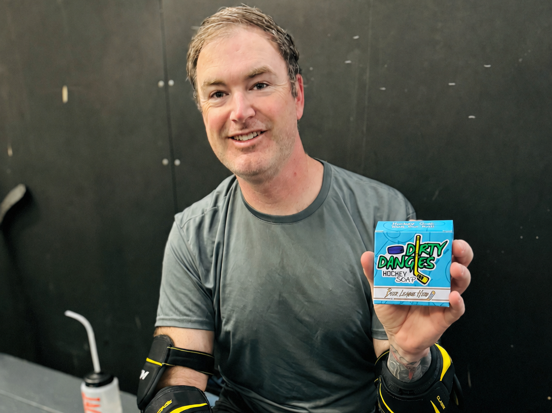 An adult beer league hockey player holding a box of league hero grapefruit pale ale soap while sitting on a bench in hockey gear