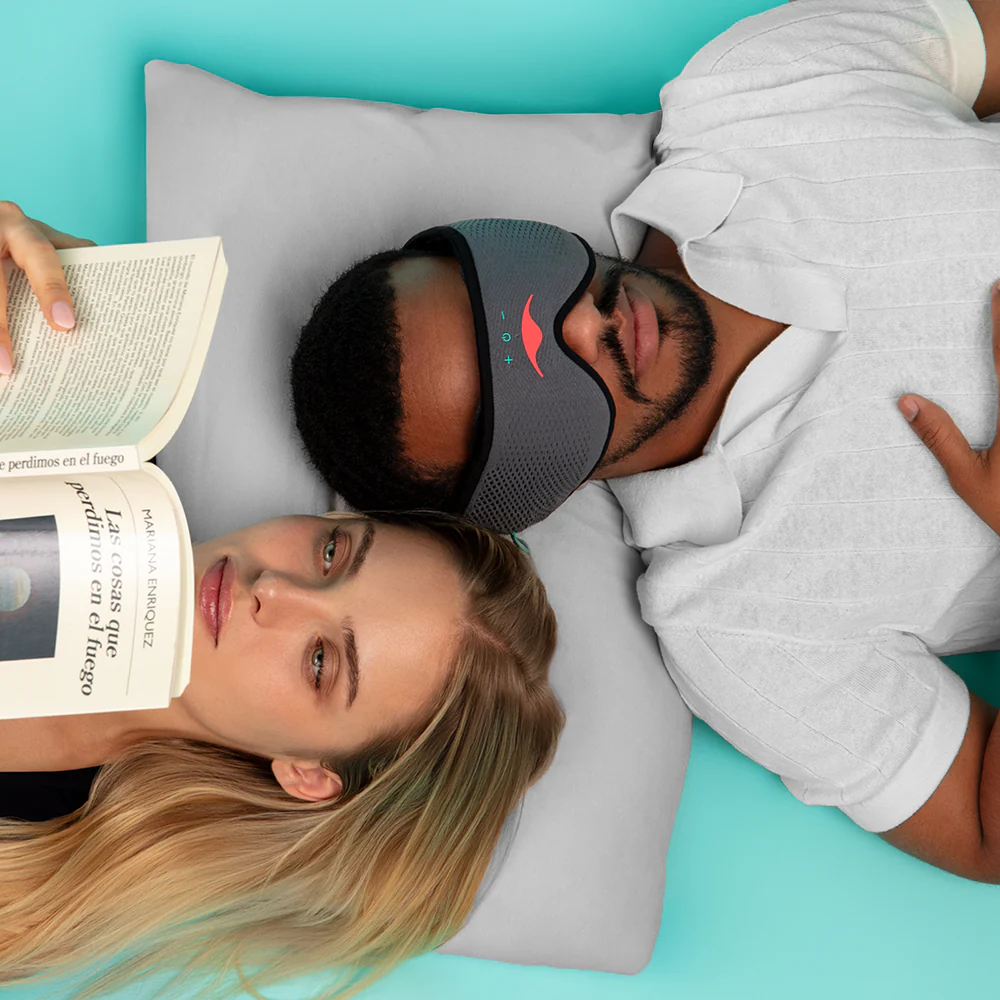 A woman and a man sharing one pillow. The man is asleep, wearing a gray Bluetooth® sleep mask, while the woman is awake and reading a book.