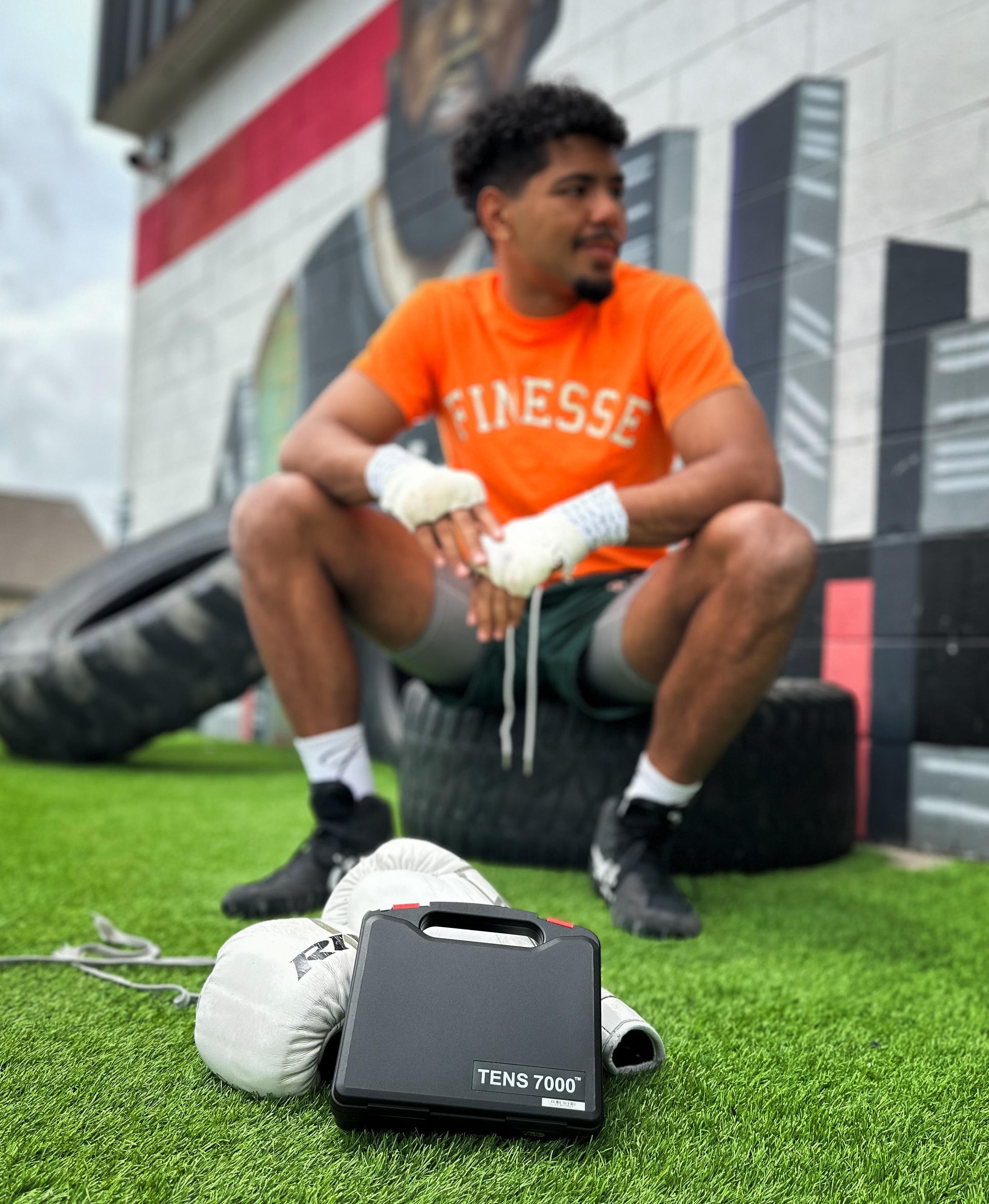 Darius sitting on a tire with a TENS 7000 device in front of him on top of boxing gloves