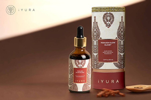 a beautiful photo of Manjish Glow Elixir - iYURA's bestselling night oil - accompanied by its beautifully illustrated, protective cylinder packaging