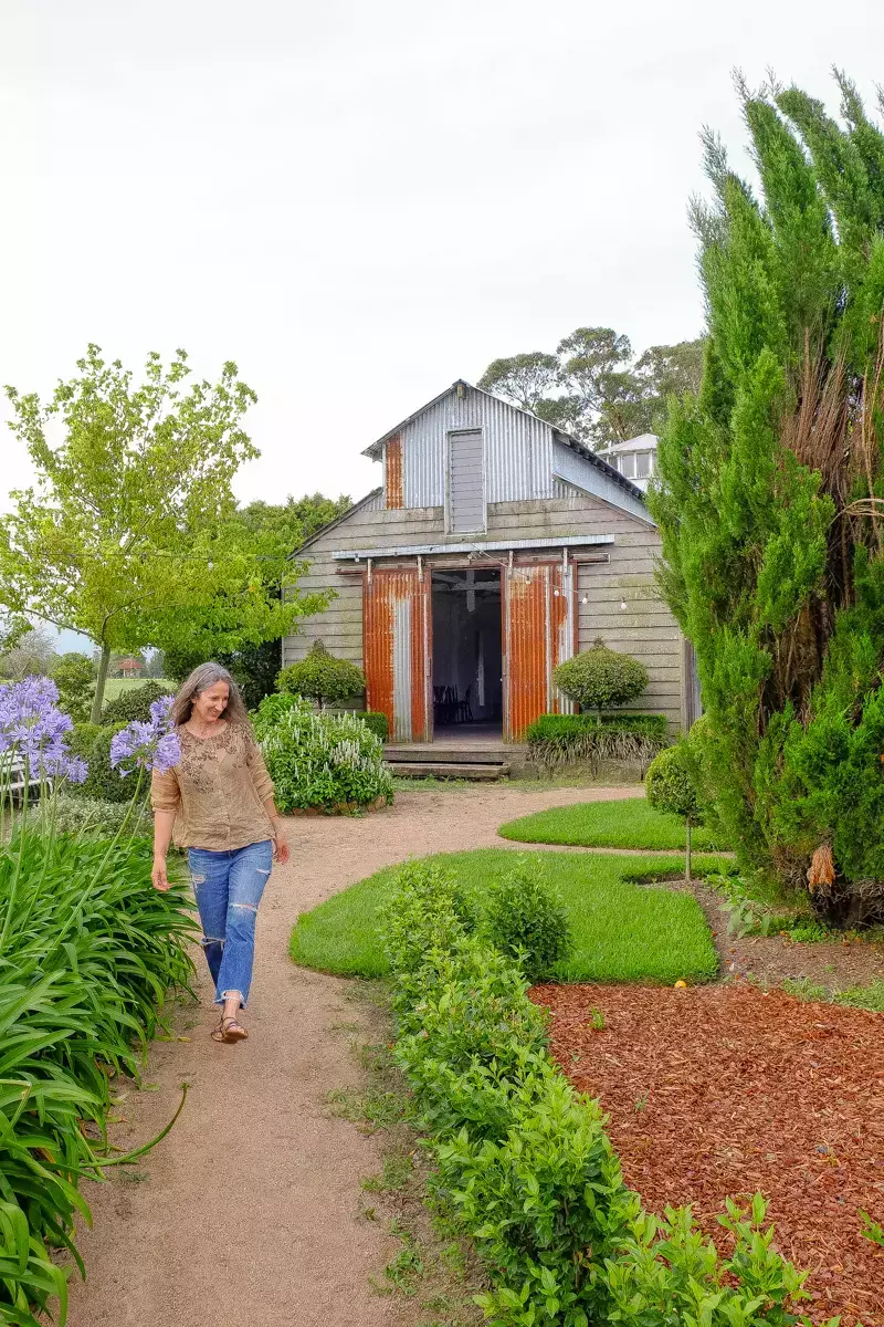 Woman walking along a curved gravel garden path toward a rustic timber barn, surrounded by manicured hedges, lush green borders, and flowering plants in a formal country garden setting.