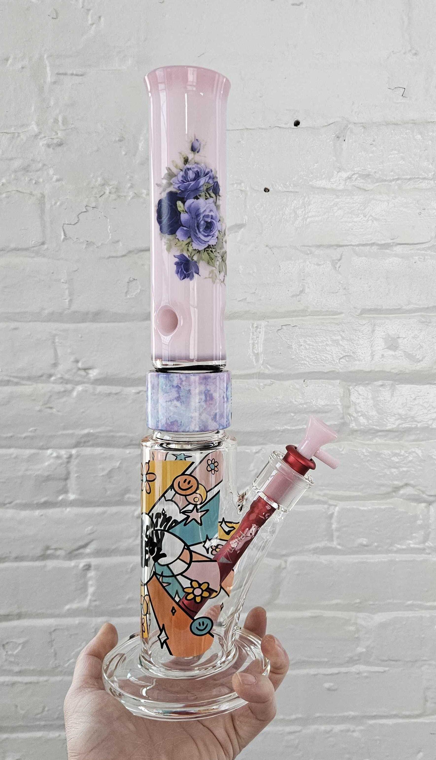 A custom Prism modular bong demonstrating "mix and match" versatility. This Single Stack features a clear Straight Tube base with the DoodleByMeg Flower Power decal, a vibrant Tie Dye Halo connector, and a solid Bubblegum Pink mouthpiece with the Midnight Rose decal. Complete with a contrast red downstem and pink bowl.