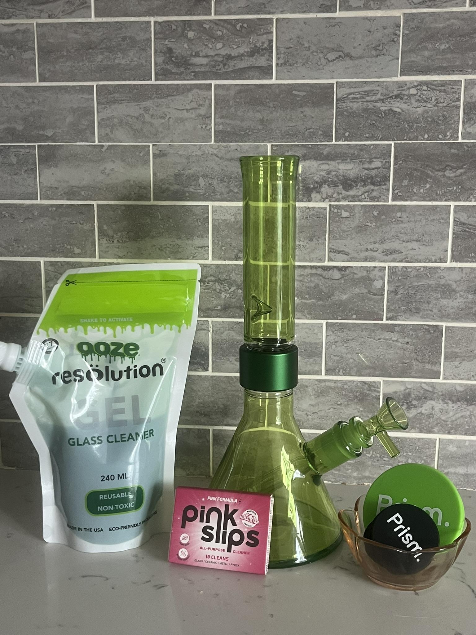 Ideal home cleaning set up in a modern kitchen, featuring a pristine, easy-to-clean Slime Green Prism MINI Monochrome Beaker Bong. The setup includes essential maintenance supplies: Pink Slips glass cleaner, Ooze Resolution Cleaning Gel, and black and green Prism Cleaning Caps, demonstrating the effortless routine required to avoid dirty bong horror stories.