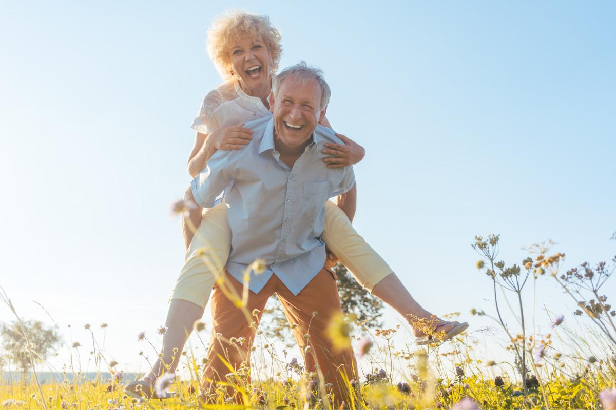 Senior woman jumping on senior man's back, smiling and laughing in the sun.