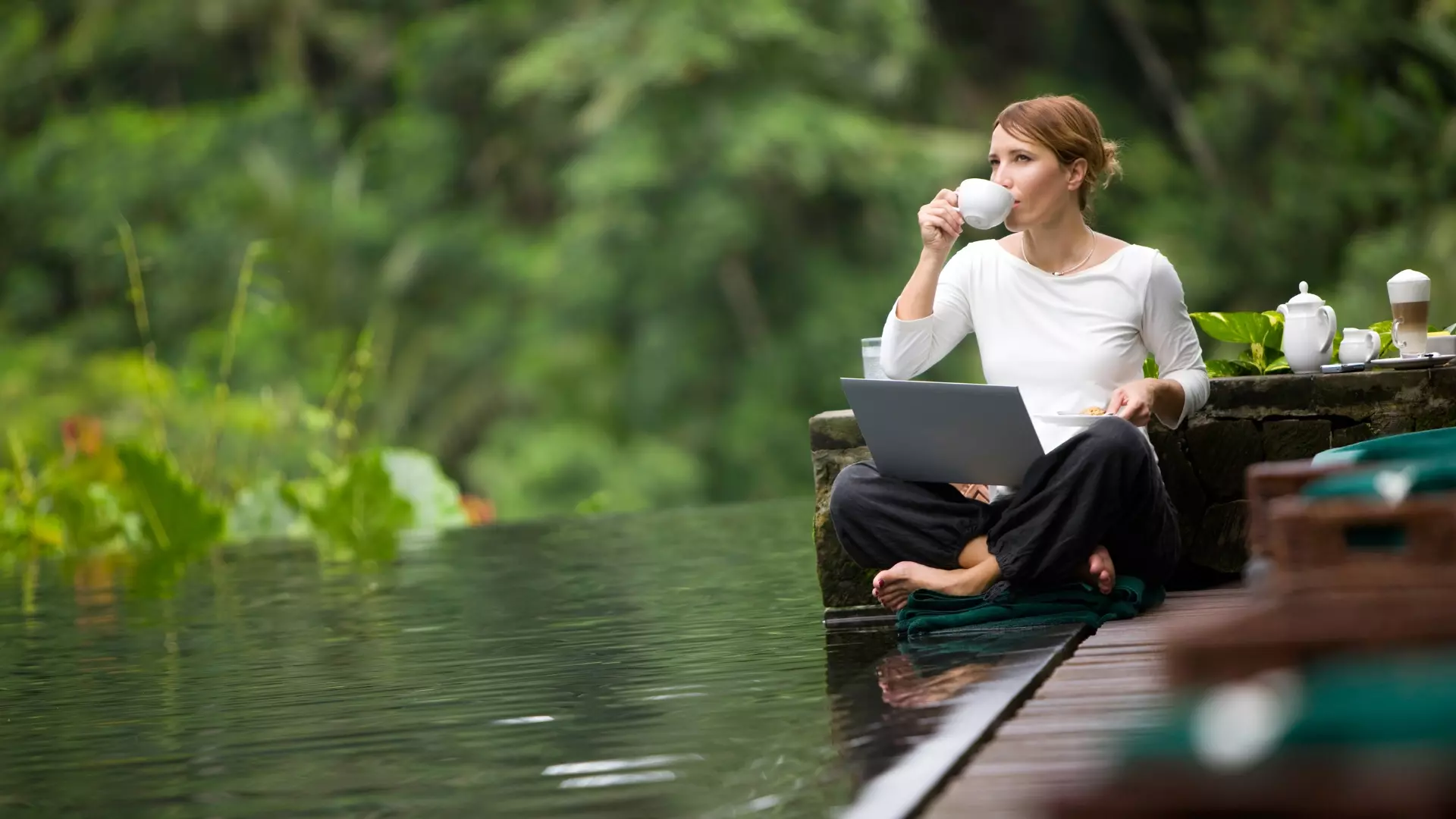 Woman drinking coffee while working on a laptop beside a calm forest pool, surrounded by lush greenery and morning light.