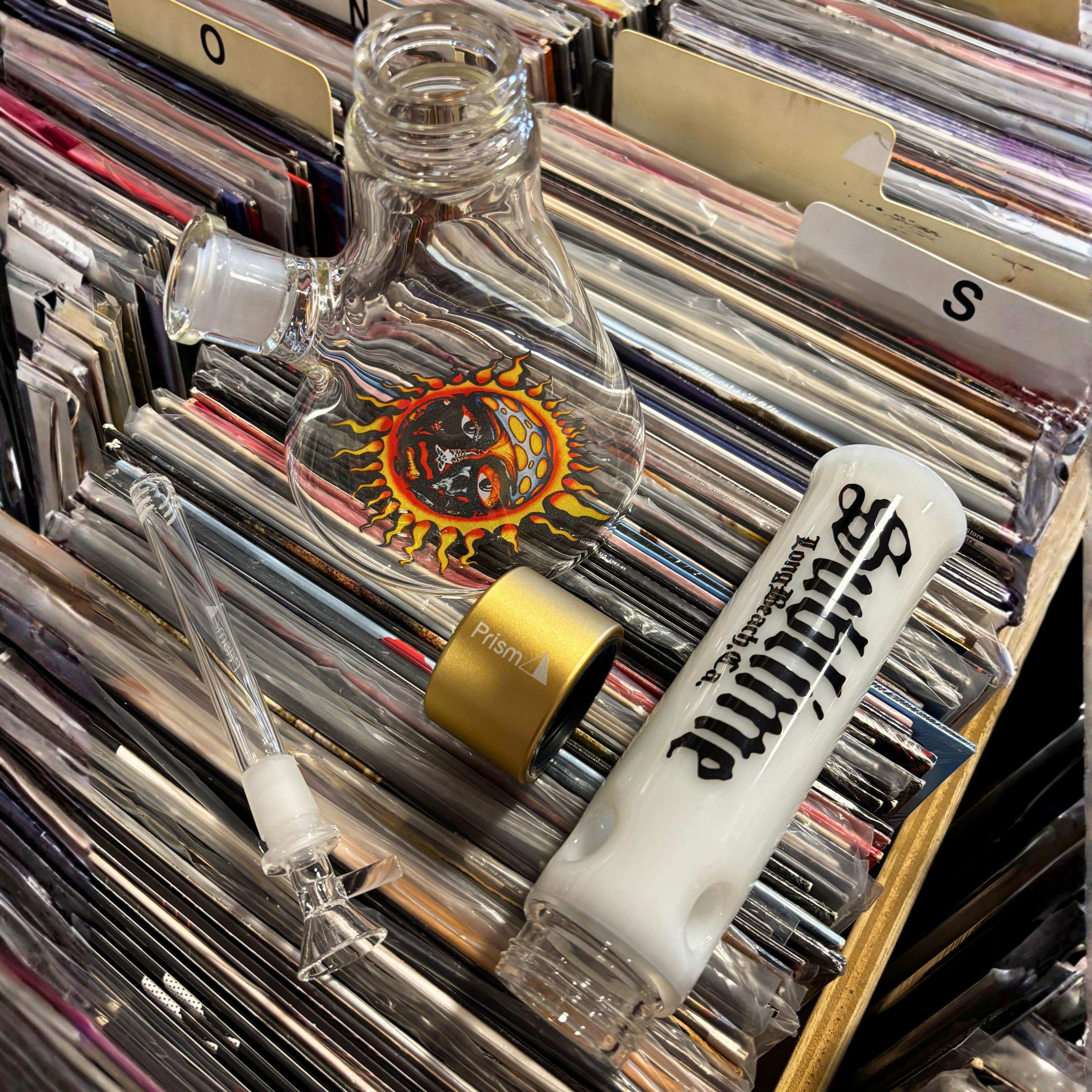 A white Sublime x Prism modular glass mouthpiece resting on a stack of vinyl records in a record store. The piece features the Gothic font reading "Long Beach, CA" as a tribute to the band's hometown and the LBC lifestyle.