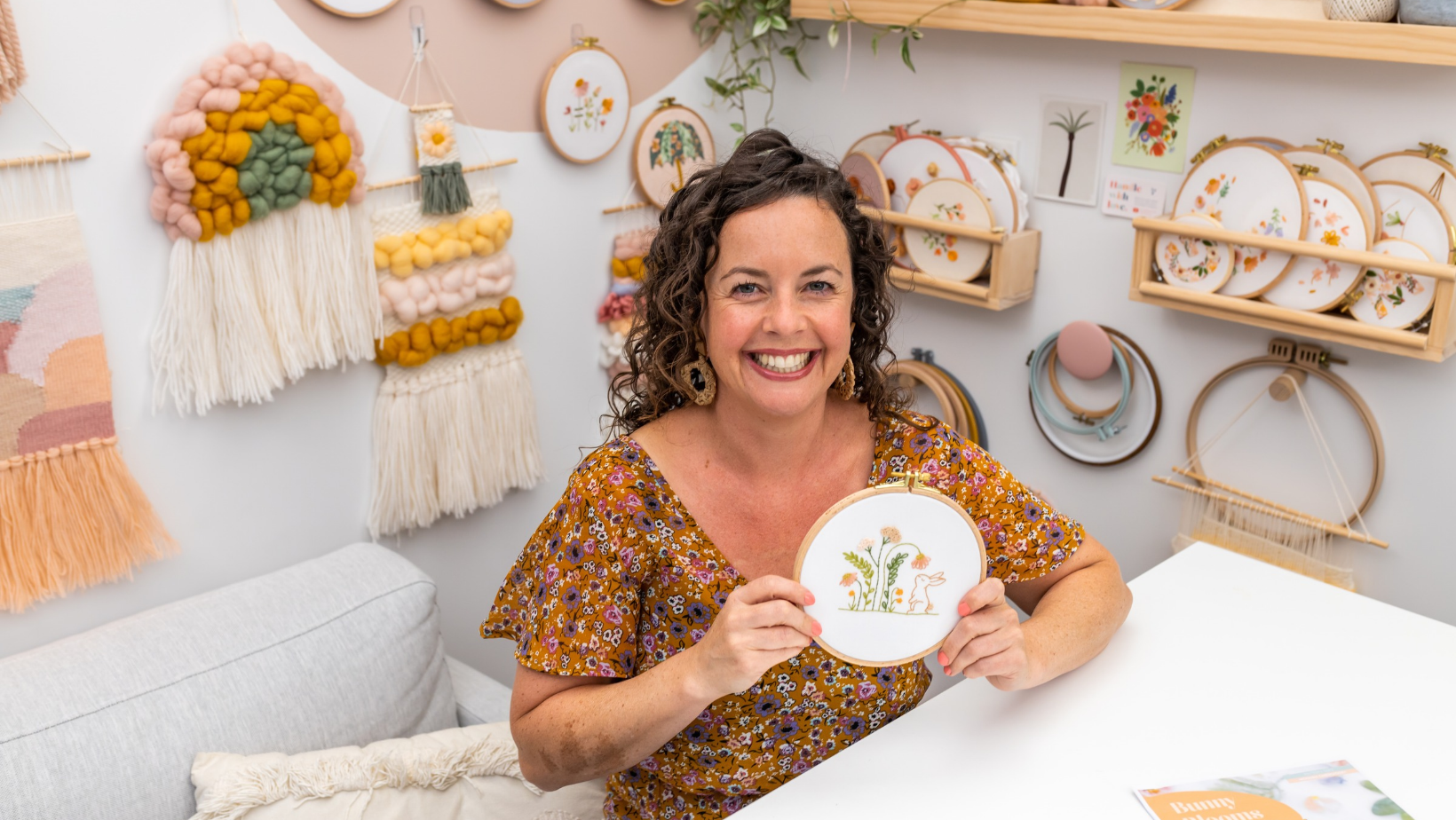 Julie Stuart holding the Bunny Blooms embroidery hoop in her craft room, surrounded by finished hoops and wall hangings.