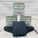 Picture of a box of Shart Wash Natural Handmade Bar Soap Deep Woods scent sitting on a black bar of soap with a wood background