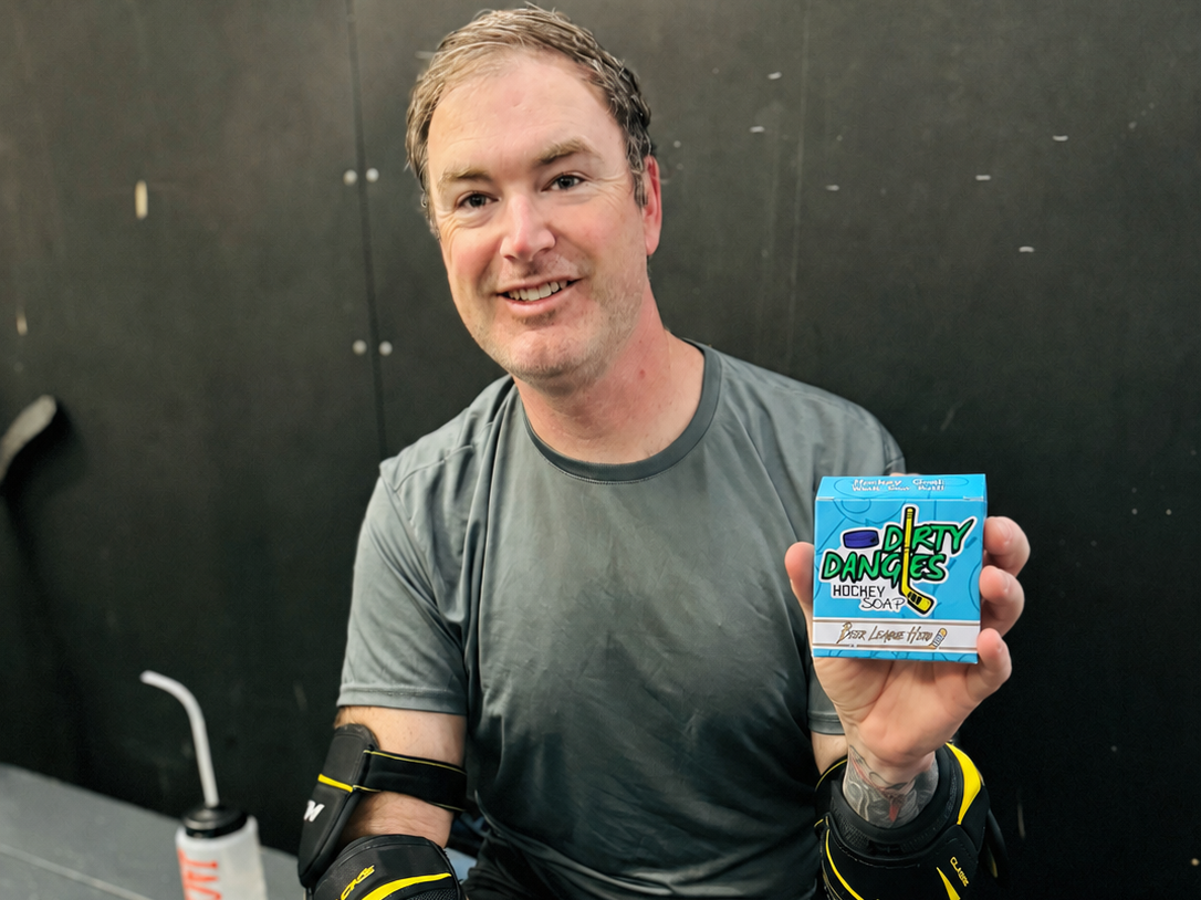 An adult beer league hockey player holding a box of league hero grapefruit pale ale soap while sitting on a bench in hockey gear
