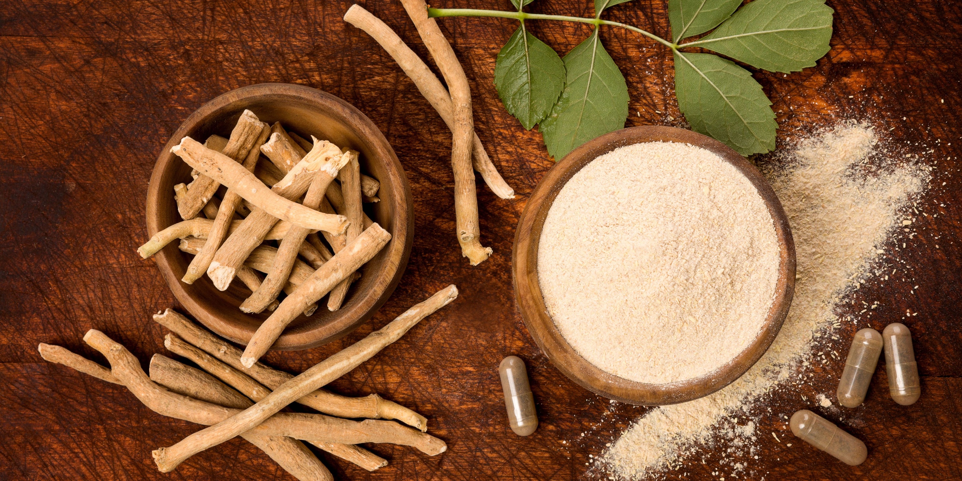 Ashwagandha roots in a wooden bowl next to powder in a wooden bowl with some capsules nearby