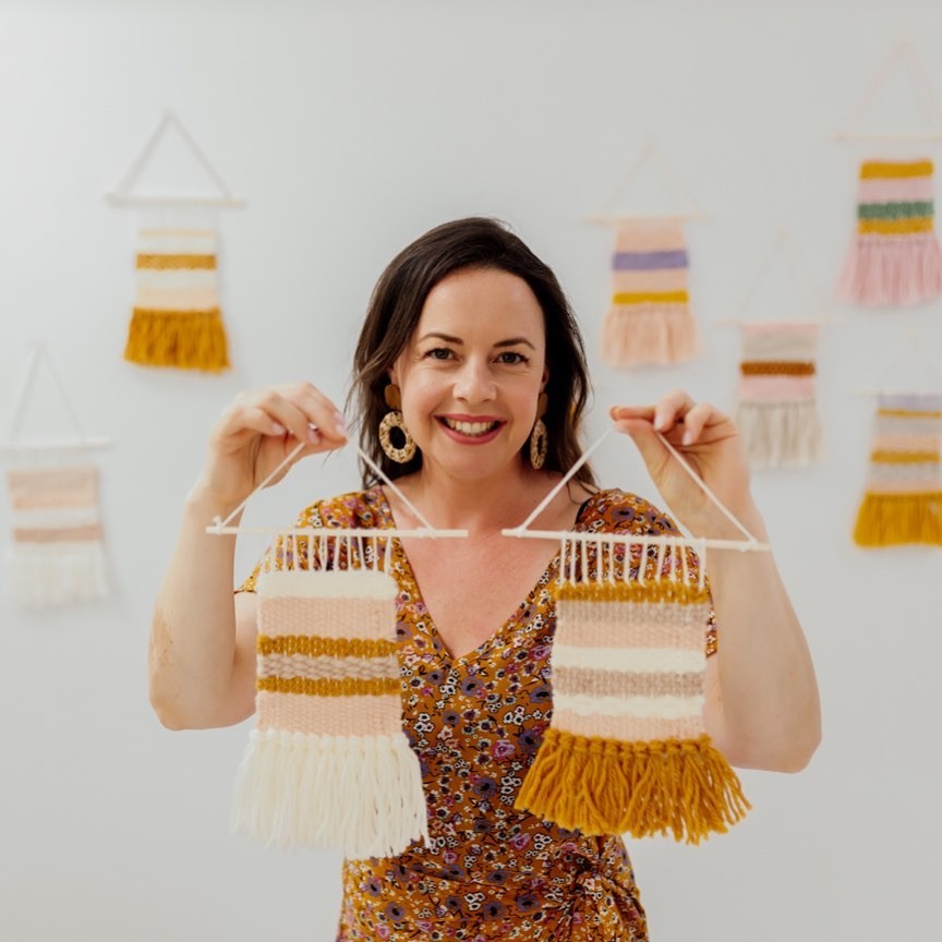 This image shows a smiling Julie holding up two mini weaves that she has woven using a DIY mini cardboard loom.