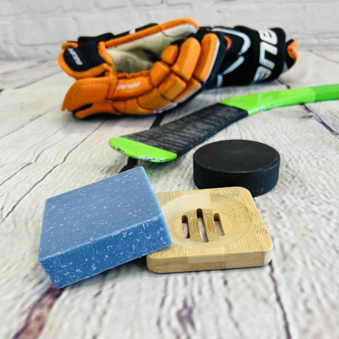 a blue soap bar sits on a bamboo soap dish sits on a wood background with a hockey stick and hockey gloves