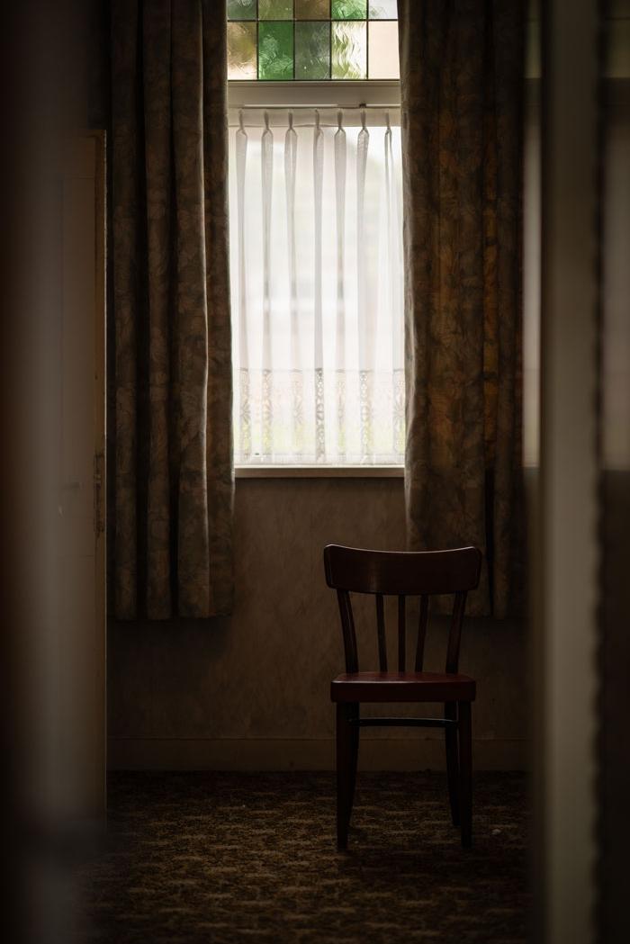 Empty wooden chair placed in a dimly lit room near a curtained window, evoking a quiet or lonely atmosphere.