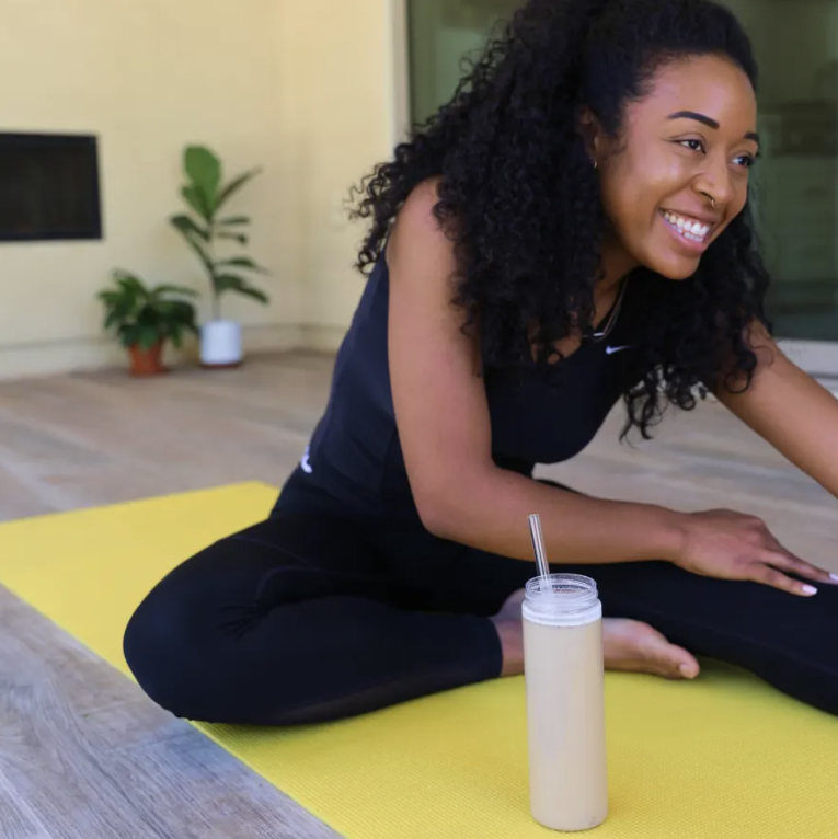 A woman sitting on a yellow yoga mat and a tall glass of white smoothie with a straw in front of her.