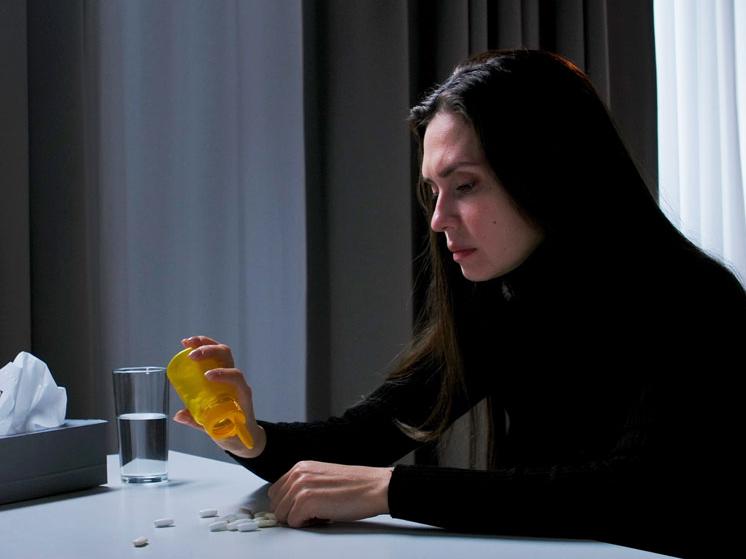 Woman sitting at a table, tilting a pill bottle as tablets spill out, with a glass of water nearby.