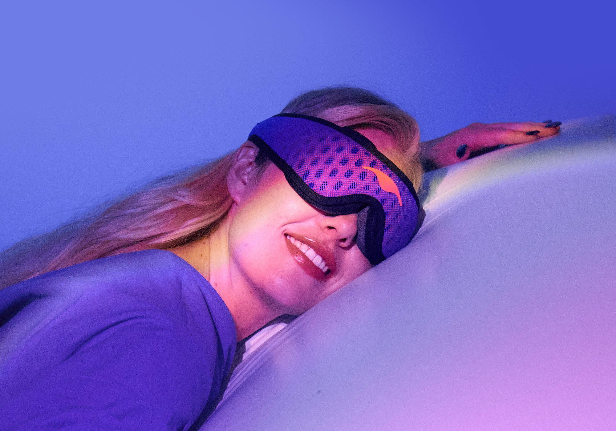 A blonde smiling girl sleeping on her side wearing an eye mask.