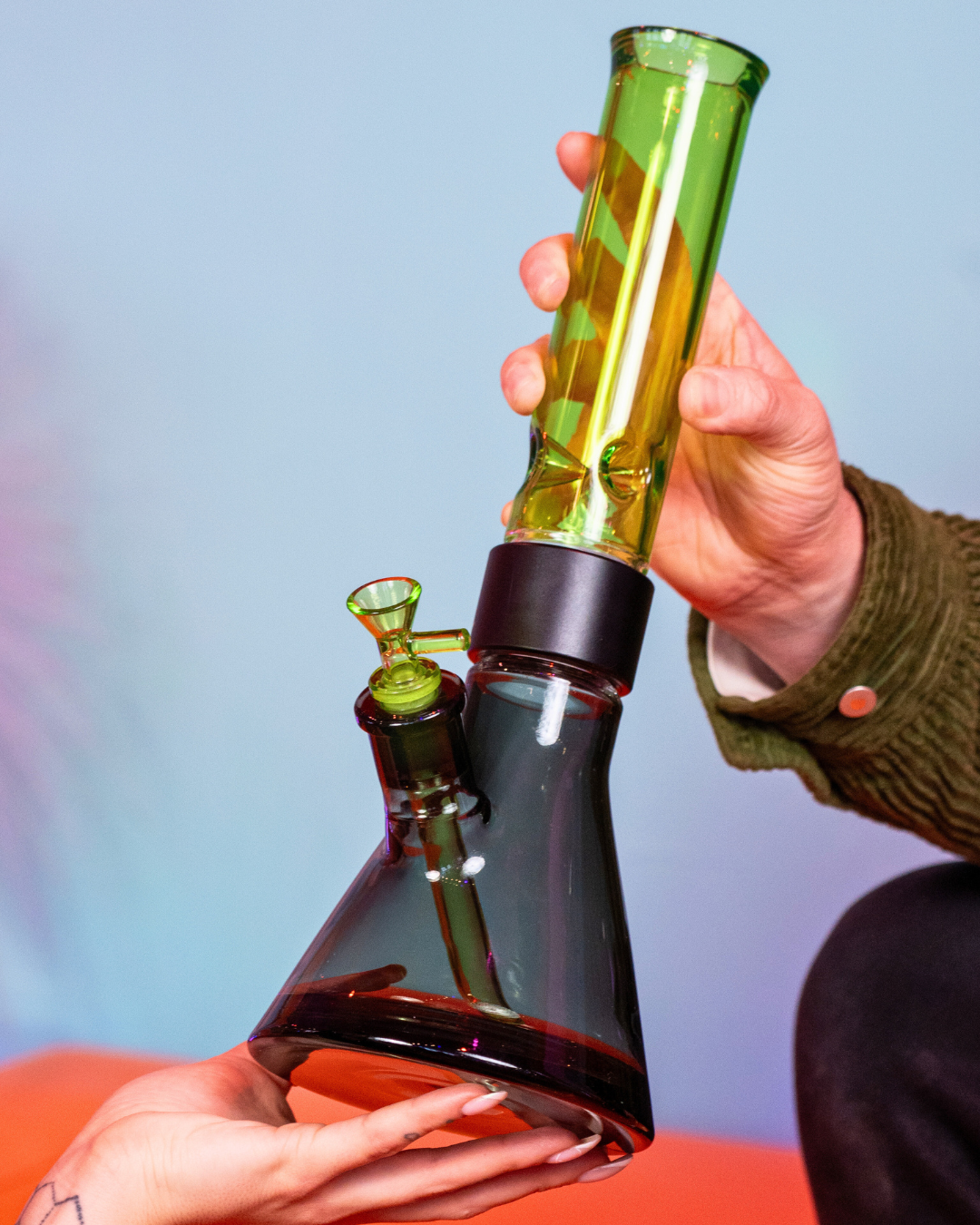 A hand holding a black and green Prism modular beaker bong, passing it to another person to demonstrate the durability and ease of use of the modular design. The image features the Slime Green glass mouthpiece, black Halo Connector, and Midnight Black base from the Duo collection.