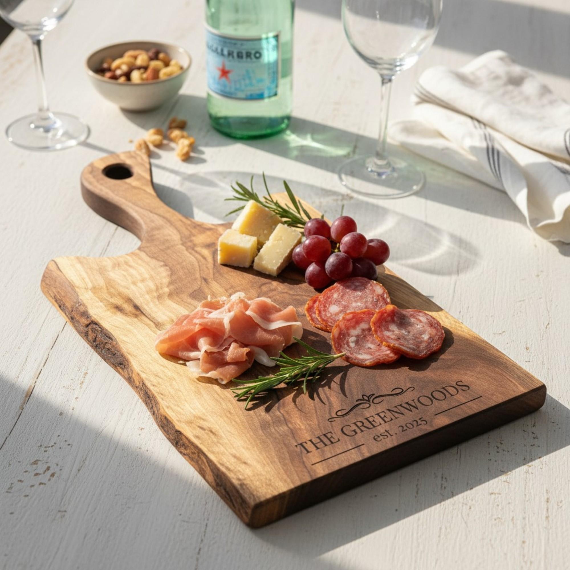 Personalized live edge walnut charcuterie board with handle carved out. styled with cured meats, cheese, grapes, and wine on a light table. perfect for housewarming and anniversary gifts