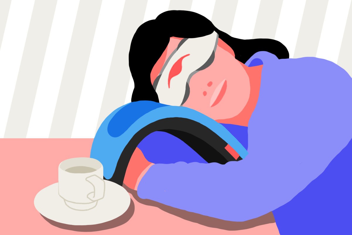 A girl taking a coffee nap on her desk, using a nap pillow and a sleep mask. There is an empty cup of coffee beside her on the table.