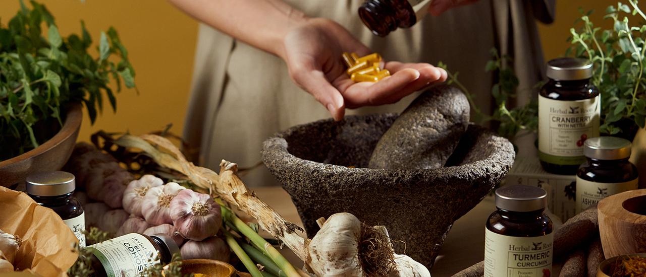 Close up of turmeric capsules being poured into an open hand above a stone mortar and pestle surrounded with garlic braids, bottles of Herbal Roots supplements, fresh basil and oregano and wooden bowls.