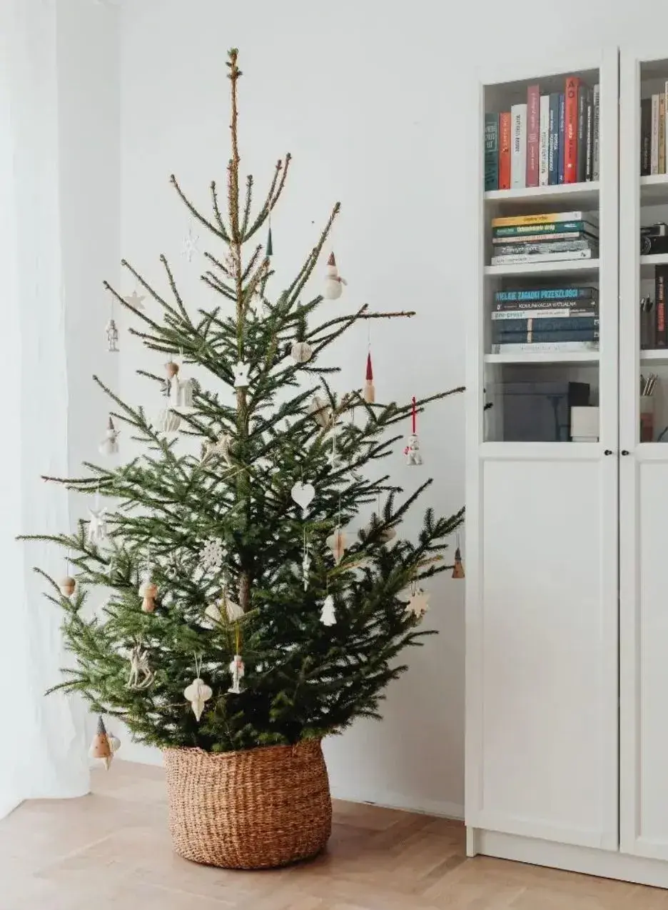A decorated Christmas tree in a woven basket, placed in a cozy room with warm lighting.