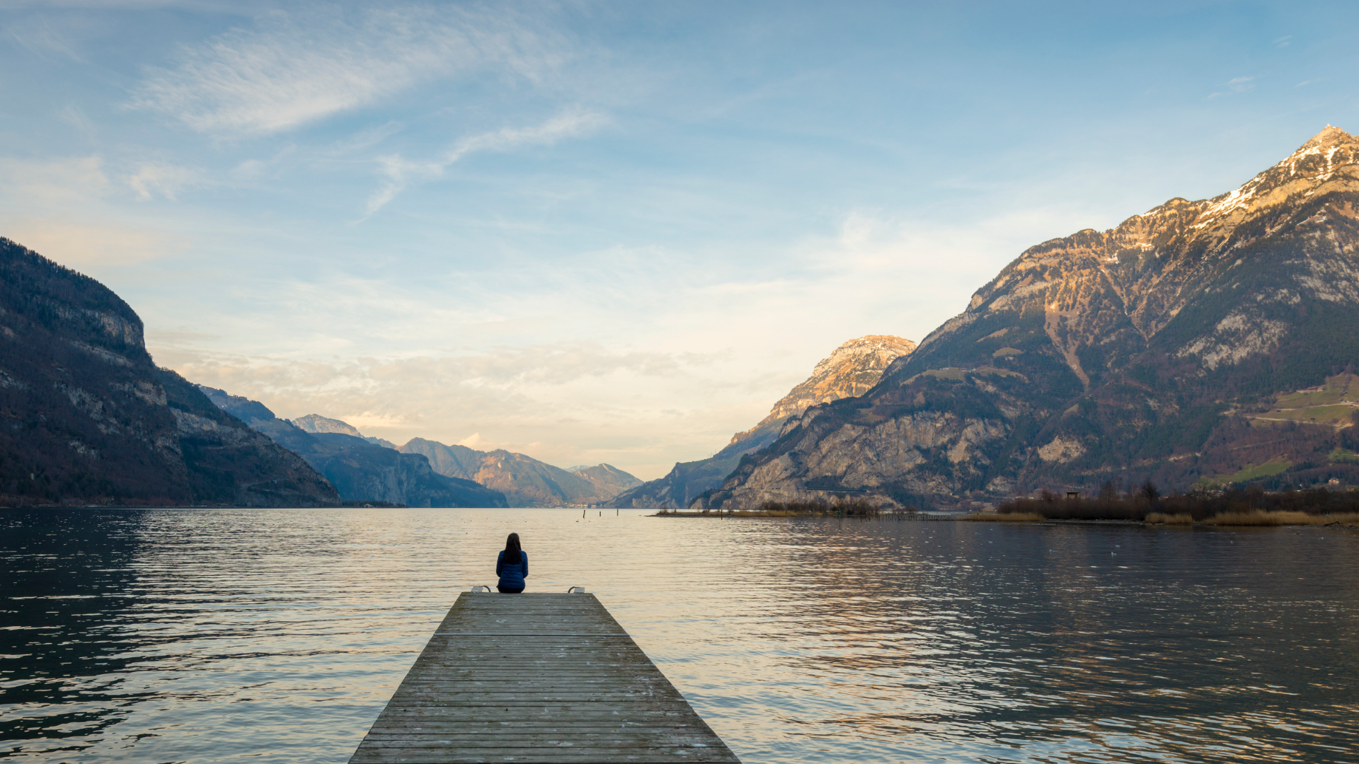 A woman sitting at the end of a pier viewing a lake and mountains at sunset.