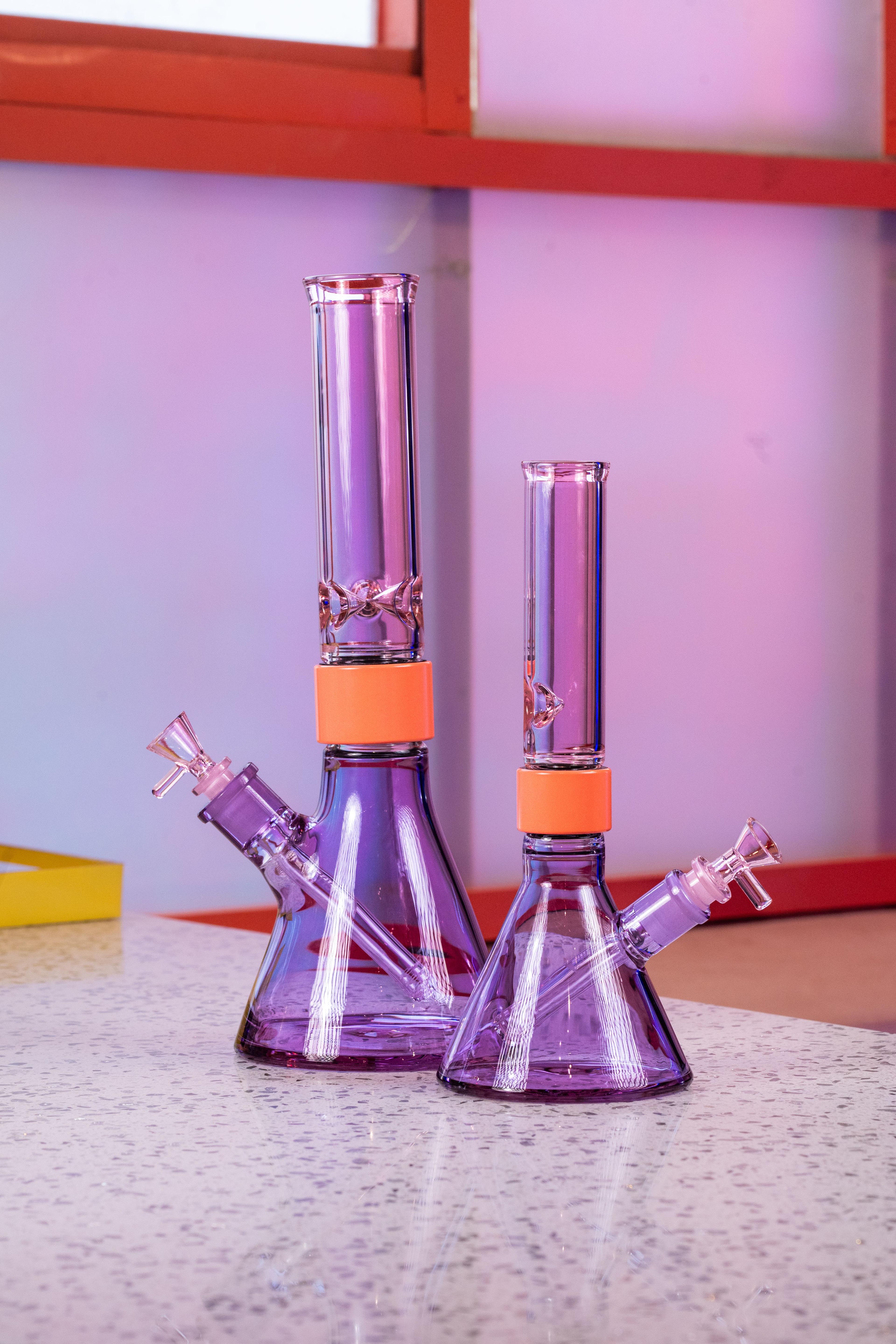 A side-by-side comparison of two pink and purple Prism modular glass bongs from the Duo Collection. On the left is the MINI size and on the right is the Classic size, both featuring standard-sized Pink Lemonade glass mouthpieces, Coral Halo Connectors, and Purple Jolly Rancher beaker bases.
